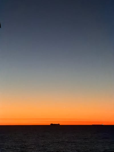 This image depicts a marine sunset or sunrise vista from City Beach, Australia. The scene is dominated by a clear horizon line separating the ocean from the sky. The sky displays a gradient of color, transitioning from a deep blue-grey at the top, through lighter blues and greys in the mid-section, to a vibrant orange and amber hue directly above the horizon. A faint light source, possibly a star or planet, is visible in the orange section of the sky, slightly to the left of the image's vertical center.

The ocean occupies the lower approximately one-fifth of the frame, appearing dark blue to black with subtle ripples indicating water movement. No distinct waves or surface features are prominently visible.

Two vessels are positioned on the horizon line. A large, dark cargo ship or tanker, appearing in silhouette, is situated slightly to the left of the image's horizontal center. It exhibits multiple lights, suggesting it is active or illuminated. Further to the right along the horizon, a second, smaller vessel is visible, also in silhouette, displaying a single, brighter light. There are no individuals, overt actions, or interactions discernible within the frame. The environment is vast and open, typical of a coastal ocean view at dawn or dusk.