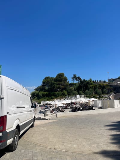 A white van is parked on a paved area at a beach resort in Crveni Vrh, Croatia. The van is facing to the right of the frame, with its rear slightly visible on the left. Beyond the paved area, a beach with numerous lounge chairs and umbrellas is set up. Several people are scattered among the lounge chairs, some appearing to be sunbathing. The beach itself is covered in pebbles. In the background, there are trees and buildings on a hillside, with a clear blue sky overhead. The scene depicts a sunny day at a resort.