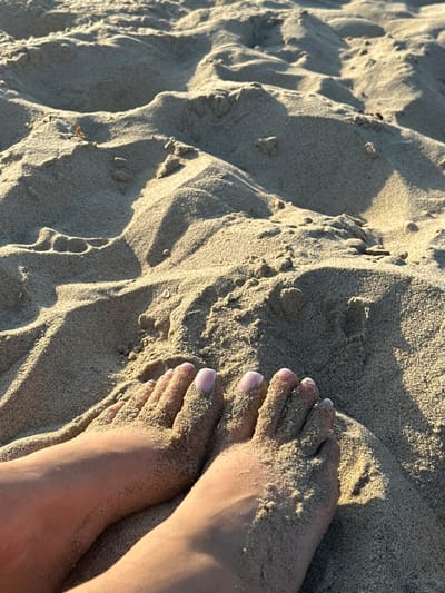 This image captures a close-up, downward-angle perspective of a pair of human feet partially embedded in light-colored sand on a beach in Manhattan Beach, United States. The feet exhibit a light skin tone, and all visible toenails are painted with a light pink or nude polish featuring white tips. The surrounding environment consists entirely of granular, light-brown sand, characterized by natural undulations, ripples, and small mounds. Strong illumination from an unseen light source, likely the sun, creates pronounced shadows across the textured sand, indicating a clear, bright day. No other individuals, objects, or structures are visible. The feet appear to be at rest in the sand.