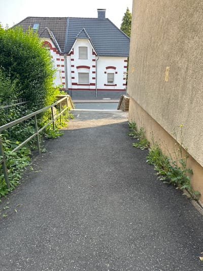The image depicts a downward sloping asphalt pathway in the city of Velbert, Germany. To the left, a metal railing separates the path from a dense green hedge. In the background, a two-story house with a dark tiled roof and white walls accented with red brick-like patterns is visible. The house has multiple windows with closed blinds. To the right of the pathway, a textured, beige-colored building wall rises. Small yellow wildflowers and green weeds grow along the base of this wall. The pathway leads towards a street or intersection at the bottom of the slope. The scene is captured in daylight with shadows suggesting late afternoon sun. There are no people or animals present.