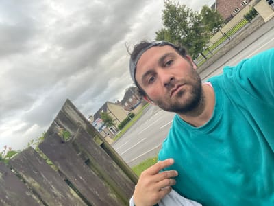 A man, wearing a teal sweatshirt and a grey headband, stands outdoors with a wooden fence and a street in the background. The sky is overcast. Houses and trees are visible behind the fence. The image appears to be taken in Armagh, United Kingdom.