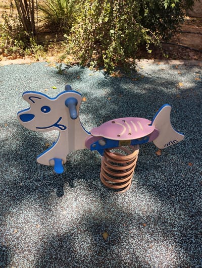 A brightly colored, child-sized spring rider is the central object in this medium shot. The rider is designed to look like a combination of a dog and a fish. The front of the rider is a stylized dog's head with blue ears and blue accents around its eyes and mouth, painted on a white background. It has blue handles for a child to grip. Attached to the dog's body is a pink, fish-shaped seat with white stripes. The fish's tail is a blue and white fin. The entire structure is supported by a large, brown metal spring. The rider is positioned on a rubberized playground surface that is speckled with blue and black. The background features dense green foliage and some dry leaves scattered on the ground. Sunlight casts dappled shadows on the playground surface, suggesting it is daytime. There is a faint shadow of what could be a tree or a structure above. The overall setting appears to be an outdoor playground, likely in a park or recreational area, given the natural foliage and the design of the play equipment. No people are visible in the frame. There is some Hebrew text visible on a small plaque on the underside of the fish seat, but it is too small and blurry to read clearly. The location is Zikhron Yaakov, Israel.