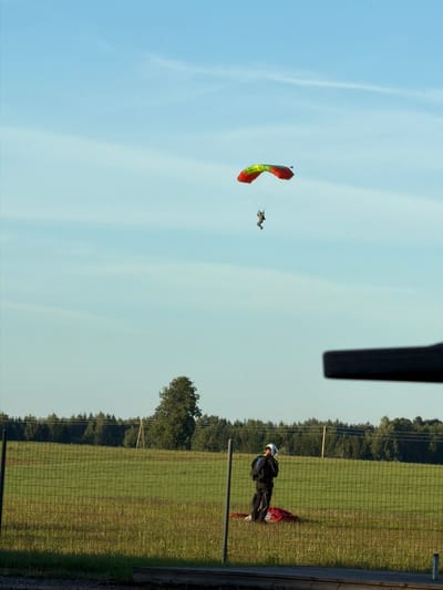 In the sky above a grassy field in Limbaži, Latvia, a skydiver descends under a red and yellow parachute. The skydiver is a small, dark figure suspended by the parachute. On the ground, behind a wire fence, two people are visible. One person, wearing a white helmet and a black jumpsuit, stands facing away from the camera, observing the skydiver. A red parachute is partially visible on the ground next to them. In the background, a dense line of trees borders the field, under a clear blue sky with sparse white clouds. A large, dark object, possibly part of an aircraft, is in the foreground on the right, obscuring a portion of the sky. The overall scene depicts an outdoor activity, likely a skydiving event, in a rural setting.