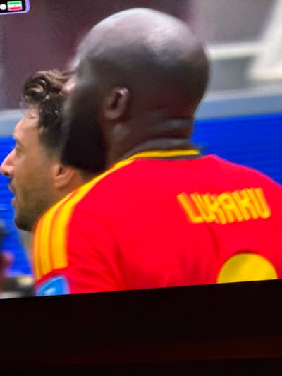 The image shows a close-up view of two men in athletic attire, likely a soccer match, with the focus on the back of one player's jersey. The player in the foreground is wearing a red jersey with yellow trim on the collar and shoulders. The name "LUKAKU" is visible in yellow lettering across the back of his jersey, along with a yellow circular emblem on the lower back. He has a shaved head. To his left, partially visible, is another man in a similar red and yellow jersey, facing left and looking away from the camera. The background is blurred, showing what appears to be a stadium environment with blue elements. There is a small flag with red, white, and green stripes in the top left corner, which could indicate a national team or event. The image is a still from a screen, possibly a television or monitor, with a visible dark border at the bottom. The scene is not located in Bykovo, Russia.