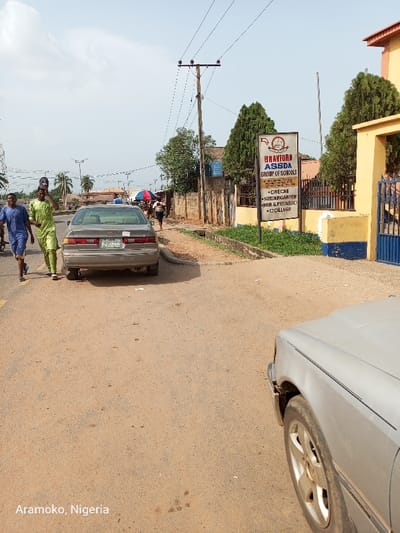 The image shows a street scene in Ado Ekiti, Nigeria, taken near the coordinates 7.656 latitude and 5.23 longitude. The scene features a paved road with a dirt shoulder, a gray sedan parked on the right side of the road, and a sign for "Bravford Assda Group of Schools" attached to a yellow building on the right side of the road. Three people are walking along the road on the left side, one is carrying a bag and the other two are wearing traditional African clothing. The scene is taken from the point of view of a person standing in the middle of the road. The image has been cropped on the right side, showing only part of a second, silver car parked on the right side of the road. The sky is clear and blue. The road is dusty and unpaved, with vegetation on the right side and some trees visible in the distance. There are utility poles on the left side of the road, with power lines running overhead. The image appears to have been taken on a sunny day with no clouds in the sky.