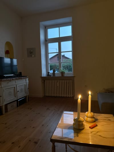 The image depicts a dimly lit interior scene, likely a living room, with a large window in the background. Outside the window, a brick house and trees are visible against a twilight sky. Inside, a white radiator is positioned beneath the window. Two lit candles in yellow and white patterned holders stand on a marble-topped coffee table, next to a red lighter. To the left of the window is a white dresser with closed cabinets, and a black television set is placed on top of it. A framed picture hangs on the wall above the dresser. The floor is made of wooden planks. The scene is consistent with a residence in Ammeldingen bei Neuerburg, Germany, given the architectural style and typical residential setting.
