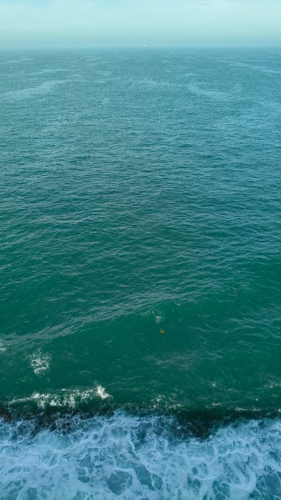 An aerial view captures the vast expanse of the sea under a hazy sky. The water transitions from a deep teal in the foreground to a lighter, almost turquoise hue towards the horizon, where a faint, distant ship is visible. White waves crash against a dark, rocky shoreline in the lower portion of the image, creating a foamy border. Several small, light-colored objects, possibly debris or marine life, are scattered across the water's surface. The scene is devoid of human presence. The location is identified as Unknown, United Kingdom.