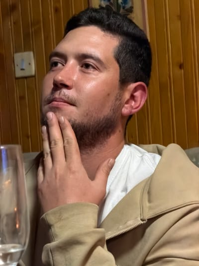 A man with dark hair and a light complexion is seen from the chest up, angled to the left. He is wearing a white undershirt and a tan hooded sweatshirt. His left hand is touching his face near his mouth. The background includes a light switch on a wooden wall. There is a clear glass object in the foreground. While the location may be Lilkovo, Bulgaria, this cannot be definitively confirmed based solely on the image.