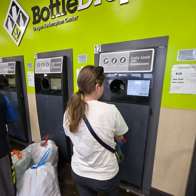 In Beaverton, Oregon, a woman with brown hair tied in a ponytail is using a bottle redemption machine. She is wearing a white t-shirt and dark pants, with a dark strap bag slung across her body. The machine, labeled with "12" and "Daily Limit 350 Containers," has a black circular opening for bottle insertion and a digital screen displaying "Sum $ 24.90" and a "Payout" button. To the left, another similar machine is partially visible. In the foreground, several large white plastic bags filled with recyclables are tied with colorful drawstrings, including a blue and a red tie. The wall behind the machines is painted a bright lime green, with signage indicating "Oregon Redemption Center" and recycling information. A beige colored wall section is visible to the right of the machines.
