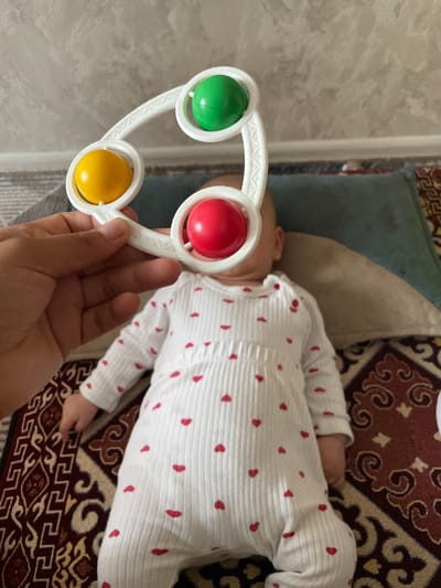 A person's hand holds a white baby rattle with three colored balls (yellow, green, and red) above a baby lying on a rug. The baby is wearing a white ribbed onesie with small red hearts. The background shows a wall with a textured finish and a pillow. The scene appears to be indoors, possibly in a home environment. The location in the city of Bujum, Kyrgyzstan cannot be determined from the image.