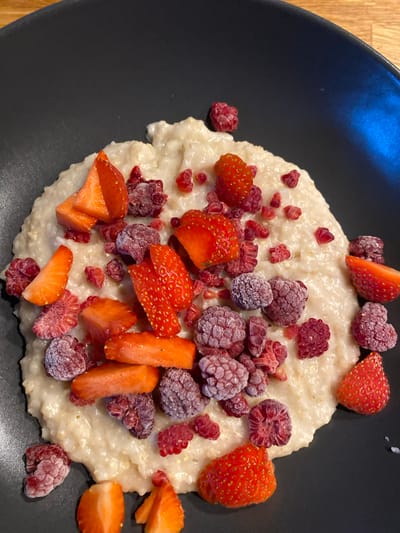 A high-angle, close-up shot shows a black plate filled with a serving of porridge topped with assorted fruits. The porridge forms a circular mound at the center of the plate, its surface appearing slightly lumpy and off-white in color. Scattered on top of the porridge are cut strawberries, varying in shape and size, with a vivid red hue. Alongside the strawberries are raspberries, some whole and some halved, many of which appear to be covered in a light frost or ice crystals. The plate sits on a wooden surface, a portion of which is visible in the lower left corner. The setting seems to be indoors, though no specific architectural details are visible. Based on the prompt, this image would be taken in Lommedalen, Norway. No people are visible in the image.