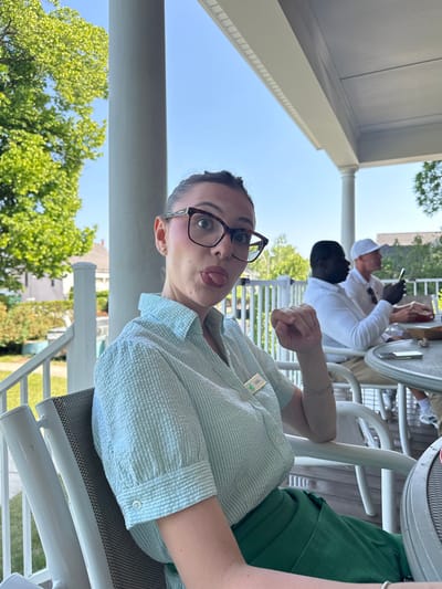 A woman with dark hair pulled back and wearing large framed glasses playfully sticks her tongue out while sitting on a porch in Harbor Springs, Michigan. She is wearing a light blue seersucker button-up shirt and a green skirt. A name tag that reads "YANA" is attached to her shirt. Behind her, two men are seated at tables. The man on the left is wearing a white shirt, and the man on the right is wearing a white cap and shirt, looking at his phone. The porch overlooks a sunny outdoor area with trees and houses in the background. The scene suggests a casual outdoor gathering or meal.