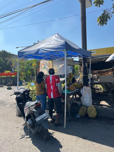 The image depicts a street scene in Sumokali, Indonesia, under a clear blue sky. A blue and white tent is set up, likely for a small roadside business. Under the tent, a woman wearing a yellow hijab is seated behind a table, which displays two large jackfruits. To her right, a white sack is visible. In front of the table, a variety of items are spread out.

Two individuals stand near the table. One, wearing a brown top and blue jeans, is facing away from the camera. The other, wearing a red and white jacket with the number "4" prominently displayed on the back, is also facing away. A black motorcycle is parked to the left of the tent, its license plate reading "W 5223 VM 06.28".

Behind the tent, a yellow building with the partial text "OP" is visible. Further in the background, trees and a sign with "RW 09" can be observed. The ground is paved with asphalt, and shadows indicate it is likely daytime. The overall impression is that of a typical street-side market or vendor stall.