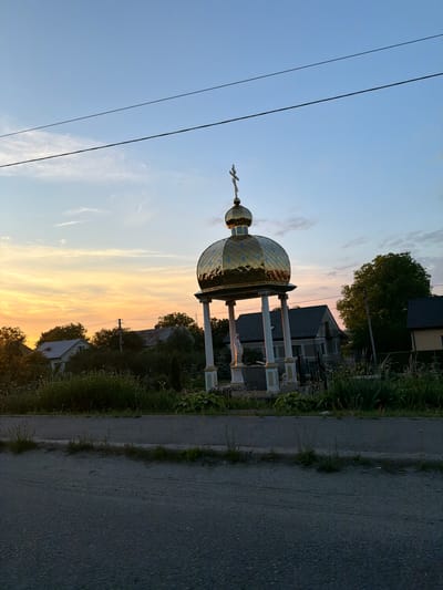 The image depicts an outdoor scene at dusk, likely in the city of Yasenivtsi, Ukraine. In the foreground, a paved road is visible, with a narrow strip of grass and vegetation separating it from a more densely vegetated area. Dominating the mid-ground is a small, ornate religious structure with a golden, domed roof topped with a cross. The structure is supported by white pillars and features a statue of a figure, possibly a religious icon, at its base. Behind the structure, several houses with dark roofs are nestled amongst trees and foliage. The sky is a gradient of blue and orange, indicative of sunset, with faint wispy clouds. Power lines are stretched across the upper portion of the frame, and trees line the background, suggesting a semi-rural or suburban environment. The overall impression is one of a quiet evening in a village or small town setting.
