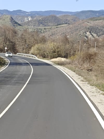 A two-lane asphalt road, marked with a solid white line on the right edge and a dashed white center line, curves through a rural, mountainous landscape in the vicinity of Gospodintsi, Bulgaria. The road initially bends sharply to the left, where a white four-door sedan is observed driving towards the viewer. A metal guardrail borders the left side of the road on this curve, accompanied by a triangular warning sign on a pole. The road surface exhibits dark tire marks and appears recently paved or well-maintained.
To the immediate right of the road is a narrow gravel shoulder, transitioning to dry grass and sparse, leafless deciduous vegetation, consistent with a late autumn or early spring season. Further along the right embankment, a pile of light-colored earth or sand is visible, along with two thin white poles. The background consists of multiple layers of undulating hills and mountains, covered predominantly by leafless trees and some patches of lighter vegetation. The sky overhead is clear and light blue. Sunlight casts long shadows across the road from the left, indicating a low sun angle. No individuals are present in the visible scene.