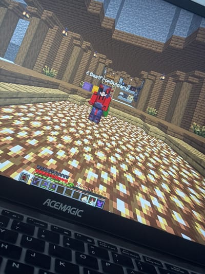 A black ACEMAGIC laptop displays the video game Minecraft, with a player character named "BestFromBulgaria" standing on a floor of alternating glowing and brown blocks. The character wears a red, black, and white outfit. The in-game environment features a large interior structure built from wooden planks and logs, illuminated by hanging lanterns. The game's user interface, including full health and hunger bars, an experience level of 38, and a hotbar displaying tools and items, is visible at the bottom of the screen. The laptop's keyboard, partially visible below the screen, includes keys with Spanish/Portuguese labels such as "Impr Pant" and "Bloq Num." This scene is observed in the city of Karnalovo, Bulgaria.