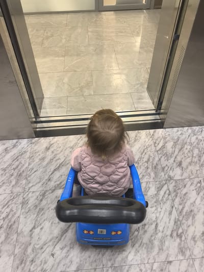 The image shows a young child from behind, sitting in a blue toy car. The child is wearing a pink quilted jacket. The toy car has a black steering wheel and a label that says "BABY CAR." The child and the toy car are on a marble-tiled floor. In the background, there appears to be an open elevator or doorway with metal framing. The scene is indoors, likely in a public space. There is no information in the image to suggest the location is Zima Juzhnaja, Russia.
