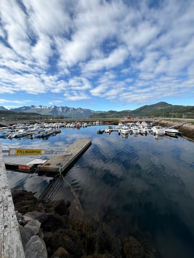 The image depicts a harbor in Ørnes, Norway, on a partly cloudy day. The foreground shows a wooden pier with a yellow sign that reads "FYLLINGSBrygge". To the right of the pier, several boats are moored in the dark, reflective water. The middle ground of the image shows a larger collection of various boats docked in the harbor, with a rocky shoreline and a road behind them. In the background, rolling green hills rise up, topped by a range of snow-capped mountains under a bright blue sky dotted with white clouds. The water's surface reflects the sky and the surrounding landscape.