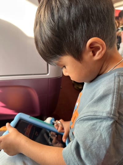 A young male child, viewed in profile from the right, with short dark hair, fair skin, and a thin silver necklace, is depicted looking down intently. He wears a light blue short-sleeved shirt with an orange graphic element visible on the right chest. The child holds a tablet enclosed in a light blue protective case, with both hands positioned on the sides. A white and blue pacifier or teether, attached by a silver ring, hangs from the top right edge of the tablet case. The tablet screen is active, displaying content that includes red and orange elements and partially visible numbers, possibly "1848". The background features a light grey or white panel with a distinct purple lower section, consistent with the interior design of modern train seating or partitions. A dark reddish-brown floor or carpet is visible beneath this structure. Blurred figures or objects are faintly visible in the deeper background to the right. The scene is located inside a train operating within the city of Breda, Netherlands.