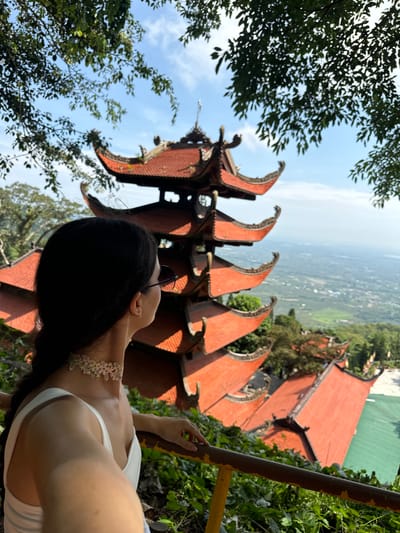 A woman with dark hair, wearing a white top and a floral choker, is captured from behind, looking out at a scenic view. Her sunglasses sit on her nose as she gazes towards a multi-tiered pagoda with red, curved tile roofs. The pagoda, a significant architectural feature, stands against a backdrop of lush greenery and a distant, hazy landscape. Trees with green leaves frame the top and sides of the image, suggesting an elevated viewpoint. The scene appears to be at a temple or cultural site, possibly in the vicinity of Thuận Nam, Vietnam, given the architectural style and the surrounding natural environment. The woman is leaning on a railing, which is partially visible in the foreground. The lighting indicates a bright, sunny day.
