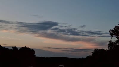 The image captures a twilight scene with a serene sky filled with wispy clouds tinged with pink and orange hues. The horizon is silhouetted by a line of trees and undulating hills, suggesting a rural or suburban landscape. In the foreground, the faint outlines of two vehicles are visible, indicating a road or parking area. The lighting and composition suggest a peaceful, transitional moment between day and night. The location of Razeni, Moldova is not explicitly depicted or inferable from the visual information provided.