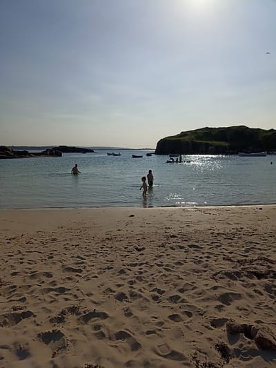 The image captures a serene beach scene at Carnboy, Ireland, under a bright, hazy sky. The foreground is dominated by a sandy beach, showing subtle undulations and scattered seaweed. Gentle waves lap at the shore, leading into clear, shallow water. Several people are visible in the water, including two young children near the shore and an adult further out. In the mid-ground, a rocky island or promontory is silhouetted against the bright sky, with a few boats anchored nearby. The water reflects the sunlight, creating a shimmering effect. The overall atmosphere is one of a peaceful summer day at the coast.