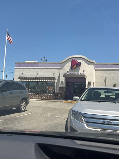 This is an eye-level, medium shot of a Taco Bell restaurant in Citrus Heights, California, on a sunny day. The restaurant has a beige stucco exterior with a purple band running along the top of the building. The Taco Bell logo, a red bell with a yellow inside, is prominently displayed above the entrance. The entrance is a dark purple, double-doorway. To the left of the entrance, a handicapped parking sign is visible. In front of the building, a silver Ford sedan is parked, facing the camera. The car has a "VETERAN" license plate frame. To the left of the Ford, a dark green SUV is partially visible, parked in a parking space. In the background, an American flag is flying on a tall white pole. Electrical wires and traffic lights are also visible in the background. The scene appears to be a drive-thru or parking lot setting.