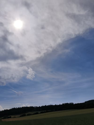 The media file displays a wide landscape shot of a rural setting on a bright, cloudy day. The sky dominates the upper portion of the image, filled with a mixture of bright white and dark grey clouds, with the sun prominently visible in the upper left corner, partially obscured by clouds and casting a bright glow. Wispy, thin clouds stretch across the sky, creating a dynamic and textured atmosphere.
Below the sky, a dense, dark green line of trees forms a treeline across the horizon. The trees appear to be on a gentle slope, with the land rising behind them. In front of the treeline, rolling fields are visible. The field closest to the trees appears to be a mix of golden-brown and green, possibly indicating harvested crops or a transition between different types of vegetation. The foreground field is a lush, deep green, with a visible texture suggesting grass or low-lying crops. A faint contrail from an aircraft is visible in the blue expanse of the sky to the left of the frame, adding a touch of human presence to the natural scene.
The overall impression is one of a serene, natural landscape bathed in the bright, diffused light of a partly cloudy day, likely late morning or early afternoon. The location is suggested to be Sebbeterode, Germany, consistent with the rural, European countryside aesthetic. No text is visible in the image. The scene evokes a sense of calm and natural beauty.