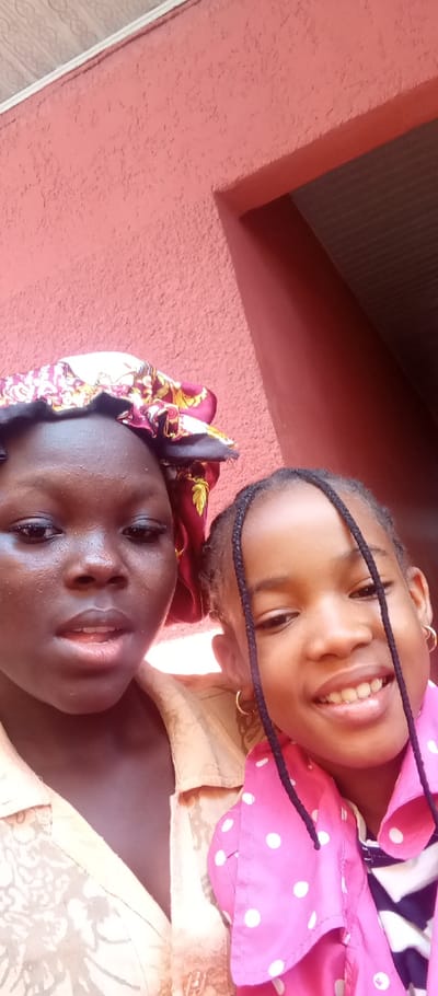 Two females are depicted in a close-up, chest-up portrait against a reddish-pink stucco-textured wall. The location is stated as Udi, Nigeria, though this is not visually verifiable from the image data.
The subject on the left is an older female with dark skin, wearing a dark red and gold patterned head covering resembling a bonnet, and a light brown or tan patterned shirt. Her mouth is slightly open, and her gaze is directed towards the camera.
The subject on the right is a younger female with lighter skin, featuring braided hair pulled back, with several braids falling forward over her shoulders. She wears small gold hoop earrings in both ears. Her attire consists of a bright pink shirt with white polka dots, worn over a dark garment with white geometric patterns. She is smiling, revealing upper teeth, and her gaze is directed towards the camera.
The background comprises the reddish-pink textured wall, with a corner or doorframe visible on the right, showing a dark, possibly brown, recessed area. A light-colored ceiling or upper wall section with a repeating pattern is visible at the top left. The subjects are positioned in close proximity, suggesting an posed interaction.