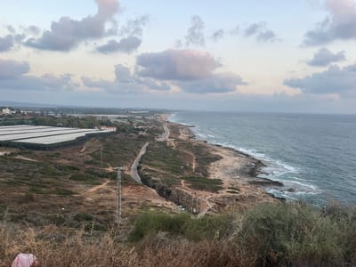 The image depicts a coastal landscape in the Mate Asher Regional Council, Israel, under a cloudy sky. In the foreground, dry brush and vegetation are visible. A dirt path winds down a hillside towards a rocky coastline where the sea meets the shore with gentle waves. A narrow road follows the coastline, with several vehicles parked along it. Further inland, a large agricultural greenhouse complex is situated on a hillside. A tall metal utility pole stands on the slope. In the distance, buildings and a town are visible along the coast. A white car is parked on a dirt path near a rocky outcrop overlooking the sea.