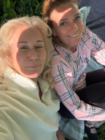 Two women are captured in a selfie outdoors, with one woman in the foreground on the left and the second woman to her right, slightly behind. The woman in the foreground has blonde, wavy hair and is wearing a cream-colored fuzzy zip-up jacket. Sunlight casts a shadow across the left side of her face. The woman behind her has reddish-blonde hair and is wearing a pink and gray plaid long-sleeved shirt over a t-shirt with a graphic design. She is smiling, and a shadow from sunlight also falls across her forehead and the upper left portion of her face. They appear to be sitting close together, possibly on a bench or outdoor seating. The background consists of green foliage, suggesting a park or garden setting. The ground beneath them is covered with a dark material, possibly gravel or asphalt, and a striped cushion is visible. The scene is illuminated by natural sunlight. The location is identified as Kerro, Russia.
