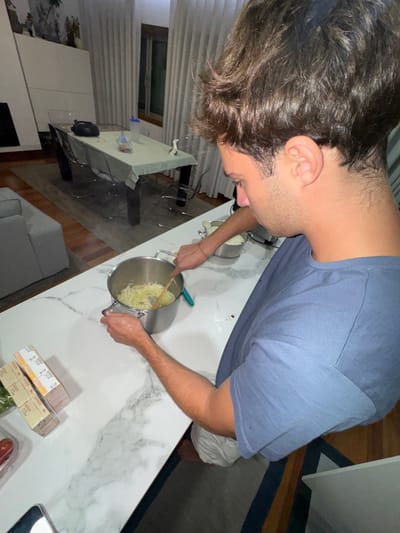 The image depicts an indoor scene within a residential property in Cidade da Maia, Portugal. A male individual, positioned with his back partially to the camera and facing left, is actively engaged in cooking at a white marble-patterned kitchen island. He has short, dark brown, wavy hair, and is wearing a plain blue short-sleeved t-shirt and light-colored shorts. His left hand grips the handle of a stainless steel pot, while his right hand uses a wooden spoon to stir the pot's contents, which appear to be thinly sliced, pale yellow food items, likely onions. A light blue-green utensil is also present in the pot. A second, identical stainless steel pot containing similar sliced ingredients is located further back on the countertop. To the left of the cooking area, a stack of four small, rectangular, light-colored packages and a clear container with green leafy vegetables and a red item are visible on the counter. A silver object is partially visible in the bottom left.
In the background, an open-plan living and dining area is visible. A dark-legged dining table, covered with a light green tablecloth, is surrounded by clear chairs. On the table, a blue water bottle, a dark electronic device, and a small white figurine are present. Further left, a section of a light grey sofa is visible. The far wall features a large, muted landscape-style wall hanging and a window with floor-length, light-colored curtains. The flooring consists of hardwood and a dark-patterned area rug. The room is illuminated by artificial lighting.