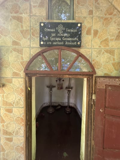 The image shows an exterior view of a small, niche-like structure with a wooden frame and a semicircular glass window above an open doorway. The walls surrounding the niche are tiled with irregular, light-colored stones. Above the tiled wall, a black rectangular plaque with white Cyrillic lettering and a cross is affixed. The plaque reads "Покой Господи. Зде лежащих. Прот. Григорія Сосновского, и его матушкѣ Леонидѣ." The doorway leads into a dark interior where two ornate, metallic candle holders stand on the floor. Between the candle holders, a small icon is visible. The floor of the interior appears to be dark and wet. A weathered red wooden door is hinged to the right of the entrance. The setting is in the village of Puchły, Poland.
