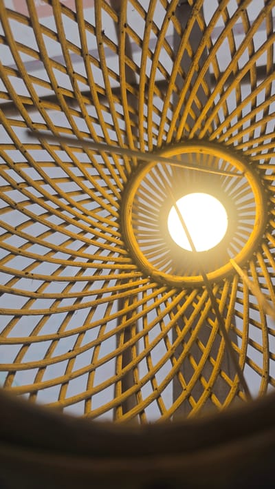 The image is a close-up, upward view of a woven, spherical lampshade with a lit bulb inside. The shade is constructed from thin, curved strands of a light-colored material, possibly bamboo or rattan, interwoven to create a mesh pattern. The light bulb at the center appears bright and white. The scene is presumably indoors. The image does not provide enough information to confirm the location in the city of Kuala Pidie, Indonesia.