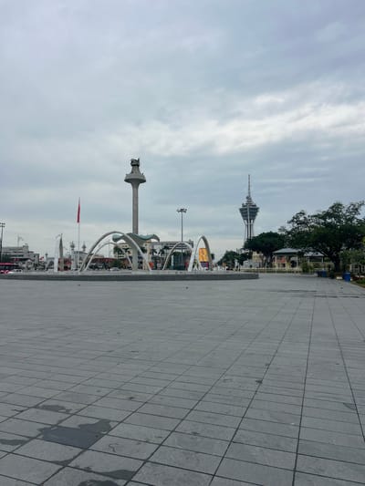 The image captures a wide, paved public square in Alor Setar, Malaysia, under an overcast sky. In the foreground, the square is tiled with large, gray square stones, some of which show patches of dampness. A low-lying, modern fountain structure with curved metallic elements dominates the mid-ground. Behind the fountain, a tall, slender monument with a distinctive top resembling an animal's head stands prominently. Further in the background, a striking observation tower with a distinctive lattice-like structure and a pointed top pierces the cloudy sky. To the right of the tower, a lush green tree provides a contrast to the urban architecture. Several buildings, including what appears to be a mosque with minarets and other low-rise structures, are visible in the distance. A red flag flies from a flagpole on the left side of the frame. A few cars and a pink bus are present on a road behind the fountain. The overall scene is quiet, with no discernible individuals in the immediate foreground or mid-ground, suggesting it might be early morning or a less busy period. The environment is urban, with a focus on public space and landmarks.