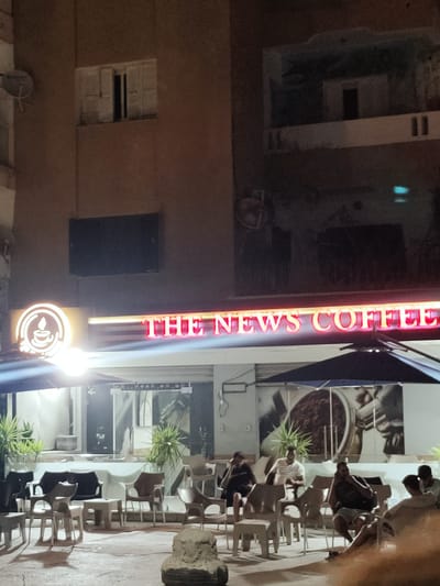 The image depicts an outdoor seating area of "THE NEWS COFFEE" establishment. The coffee shop's sign, illuminated in red neon letters, is prominently displayed across the facade. To the left of the sign is a circular logo featuring a steaming cup. A black umbrella provides shade over a section of the seating area, where several individuals are seated at tables. The setting appears to be in the city of Ariana, Tunisia. The time of day is evening or night, as indicated by the artificial lighting.