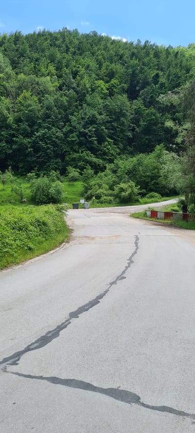 The image presents a rural landscape in Iamna, Bulgaria, under a clear blue sky with minimal cloud cover. A dense, verdant forest, primarily composed of deciduous trees, covers a prominent hillside dominating the background and extending to the lower right. In the foreground, a light gray asphalt road is visible, featuring a distinct, dark, wavy asphalt repair patch or crack running down its center towards the viewer. The road appears to fork or intersect in the middle ground. On the right side of the road, where it curves, a short barrier or railing painted with alternating red and white stripes is present. To the left of the road, near the intersection, two gray outdoor bins are situated alongside a small yellow square sign on a post and a partially obscured white sign. Lush green bushes and grass line the road's edges, particularly on the left. No individuals or animals are visible in the scene.