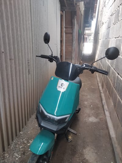 An electric scooter, colored teal, is parked in a narrow alleyway in Kedung Dowo, Indonesia. The scooter is positioned facing forward, with its handlebars and front fairing prominently visible. The scooter has a logo on its front that appears to be circular with stylized text.
The alleyway is flanked by two walls: on the left, a corrugated metal wall with vertical ridges, and on the right, a wall constructed of concrete blocks. The ground in the alley is dirt with some gravel and debris. Overhead, the alley is partially covered by structures made of wood and corrugated metal, suggesting residential or workshop environments. Light is filtering into the alley from a brighter area at the far end, indicating an opening or exit. There are no people visible in the image. The overall scene depicts a utilitarian space in a residential or semi-industrial area.