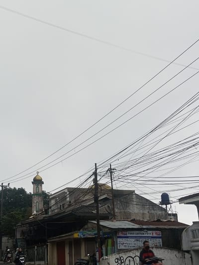 An overcast sky fills most of the frame, contrasting with the buildings clustered below. A mosque with a green and gold minaret peeks out from behind other structures. Power lines crisscross the view. Buildings vary in age and condition, some with visible wear. A water tank sits on one roof. Motorbikes are seen on the street level with one rider wearing a mask. Graffiti is visible on a wall.