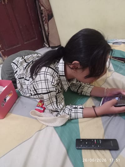 The image shows a young woman lying on a bed, playing on her smartphone. She is wearing a black and white checkered shirt and has her hair tied back. To her left, there is a pink plastic basket containing various items. In front of her, a red packet of Biskuat crackers is visible. On the bed, near her right hand, is a black remote control with various buttons and icons. The bed is covered with a patterned sheet. In the background, a wooden door and a cream-colored wall can be seen. The timestamp "26/06/2026 17:51" is overlaid on the bottom right of the image. The scene appears to be indoors, likely in a bedroom. While the image itself does not explicitly indicate the city of Sukaraja, Indonesia, this information has been provided as context for the scene.