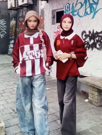 The image depicts two young females standing outdoors on a paved surface in Aktash, Uzbekistan, against a backdrop of graffiti-covered walls. The ground is composed of light-colored interlocking pavers.
The individual on the left, facing directly forward, wears a light brown hijab over a dark brown underscarf. She is dressed in a long-sleeved maroon and white striped shirt featuring a white collar and a prominent white graphic on the chest. Her lower attire consists of loose-fitting, light-wash blue jeans, with her hands placed into the pockets. A light-colored shoulder bag strap is visible over her right shoulder.
The individual on the right, positioned slightly behind and to the right of the first, wears a dark red hijab over a matching underscarf. She is dressed in a long-sleeved dark red shirt with a white collar, a small gold-colored emblem on the left chest, and white "FF" lettering on the right chest. Her lower attire consists of loose-fitting dark gray pants. Her left hand holds a light-colored smartphone, and a watch with a brown strap and gold-toned face is visible on her left wrist. A necklace with a small pendant is also visible. Her gaze is directed towards her right.
The background features two adjoining wall sections. The left section is dark-colored, with an air conditioning unit and associated piping mounted high, and is extensively covered in white and light-colored graffiti. The right section is a white-plastered wall adorned with multi-colored graffiti in black, blue, and red, and includes a small, dark-colored window secured with bars. A simple concrete bench with brick supports is situated against the wall on the right side of the frame.