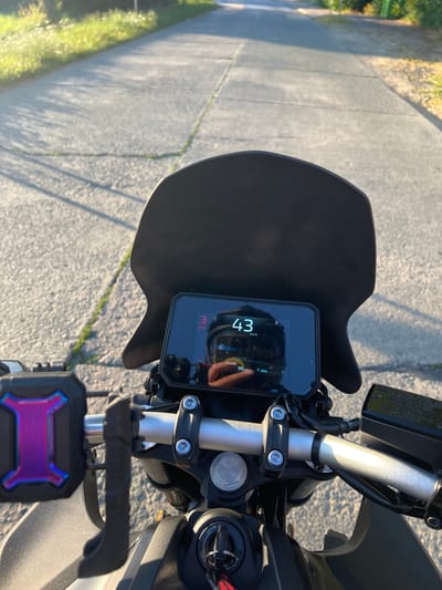 A first-person perspective, point-of-view shot of a motorcycle dashboard shows a digital display with the current speed at 43 km/h. The display also indicates a gear of "3", a time of "8:18", and a "RANGE" of 96 km. The motorcycle is on a paved road with sunlight casting shadows. The environment appears to be a rural area, possibly on the outskirts of Nieuwenrode, Belgium, as indicated by the paved road with grass and vegetation growing along the edges, under a clear sky. The handlebars, grips, and a smartphone mount with a colorful phone case are also visible. A reflection of the rider wearing a helmet can be seen on the dashboard screen.
