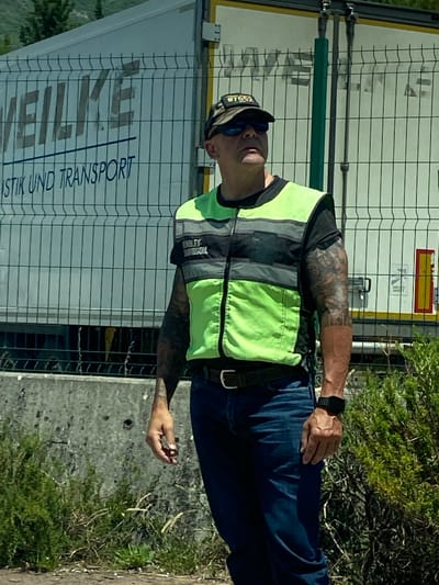 A middle-aged man wearing a neon green and gray safety vest over a black t-shirt and dark jeans stands near a chain-link fence. He has short, dark hair, sunglasses, and a camouflage baseball cap with a patch that reads "W7532". His left arm is heavily tattooed. The man is looking upwards and to his right. Behind him is a white truck with blue lettering that appears to say "WEILKE LOGISTIK UND TRANSPORT". The ground in front of the fence is a rough, gray concrete surface with some sparse vegetation. The background shows a slight incline with green trees and a hazy sky. The location is identified as Palje Brdo, Croatia.