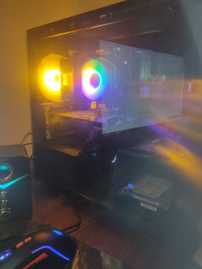 This is a close-up, angled shot of the interior of a computer tower, likely on a desk. The computer is illuminated by internal RGB lighting, with a prominent fan showing a vibrant rainbow spectrum of colors at the top, and another fan below glowing a bright yellow. The lighting creates a colorful and somewhat blurry effect on the components within the case.

Visible components include a hard drive mounted in the upper right section of the case. Below it, a motherboard is partially visible, with the text "ASUS H61M-D" clearly legible on a component. A power supply unit with the "QS 750" designation is also visible towards the right.

In the foreground, to the right of the computer tower, a gaming mouse with blue and orange LED accents is visible. To the left and below the computer, a speaker with blue and green lighting is also present. The overall scene suggests a gaming or computer enthusiast's setup. The lighting indicates it's likely nighttime or a dimly lit room. The presence of specific computer hardware components and peripherals are the main subjects. There are no people or animals visible. The background appears to be a wooden surface, possibly a desk. There are no overt environmental cues like weather or specific location identifiers, beyond the "Location context" provided, to suggest anything outside the immediate computer setup.