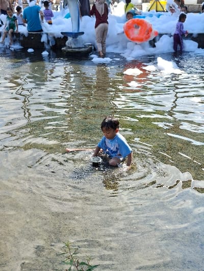 A young boy in a blue shirt is sitting in a shallow pool of water, playing with a small object. He is in a water park in Kecamatan Tulung, Indonesia, located at latitude: -7.592, longitude: 110.65. The pool is surrounded by white foam and a dolphin sculpture. The boy's feet are submerged in the water. Behind him are several adults and children enjoying a water park activity. There is a large red inflatable ring lying on the foam. The water is clear and the ground is sandy. The sun is shining brightly.