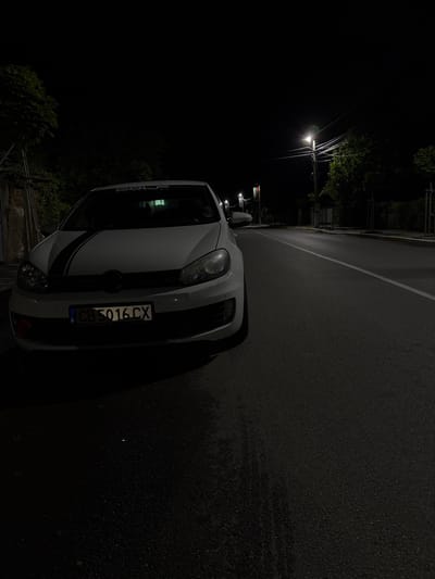 The image depicts a white car parked on a street at night in Dolna Banya, Bulgaria. The car, which appears to be a Volkswagen Golf, has a distinctive black stripe running down the center of its hood and front bumper. A sticker partially visible on the top of the windshield reads "GOLF MOTORSPORT." The front license plate displays "CB 5016 CX," with the country code "BG" for Bulgaria, and a small "B" symbol, likely indicating the registration district.
The vehicle is positioned alongside an asphalt road that appears damp or wet, reflecting the dim light available. A white solid line marks the edge of the road. In the background, a solitary, brightly lit street lamp illuminates a portion of the scene, casting light on some trees and foliage that line the left side of the street. Numerous power lines extend from the pole holding the street lamp, silhouetted against the dark night sky. No activity or people are visible; the scene focuses on the stationary car in a quiet, nocturnal setting.