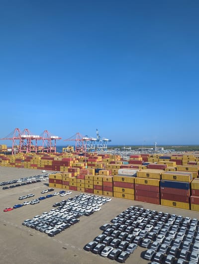 The image shows a large container terminal at the Port of Hambantota, Sri Lanka. The scene is dominated by stacks of colorful shipping containers, predominantly red and yellow, with some blue ones visible. Several large cranes, painted pink and blue, are positioned at the port, suggesting active cargo operations. In the foreground, a vast expanse of a paved surface is covered with rows of parked cars, mostly white and silver, arranged in an organized manner. The sky above is a clear, bright blue. The environment appears to be a sunny day in a busy port city.