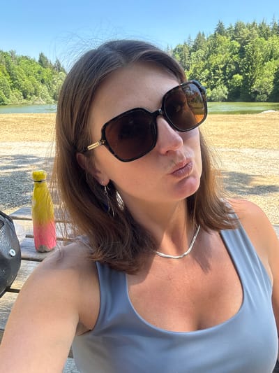 A woman is taking a selfie outdoors. She is wearing large, dark-rimmed sunglasses and a light blue tank top. She has brown hair and is wearing a thin silver necklace and long, dangling earrings. Her lips are pursed. She is sitting at a wooden picnic table, and a brightly colored water bottle is visible on the table to her left. Behind her, there is a body of water, likely a lake, with a densely wooded hillside beyond it under a clear blue sky. The area appears to be a park or recreational area.