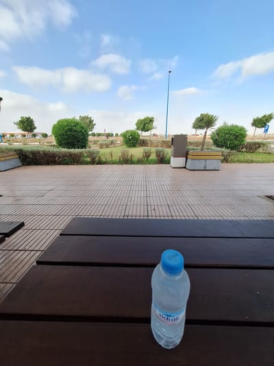 The image depicts an outdoor scene on a paved area with a wooden table in the foreground. A plastic water bottle with a blue cap is placed on the table. Beyond the paved area is a grassy expanse with various trees and shrubs, including some neatly trimmed bushes and a solitary palm-like tree. In the background, a lamppost stands tall, and further beyond, buildings and structures are visible, suggesting an urban or semi-urban environment. The sky is partly cloudy, with blue patches and white clouds. The scene appears to be in Oulad Hcine, Morocco, given the context.