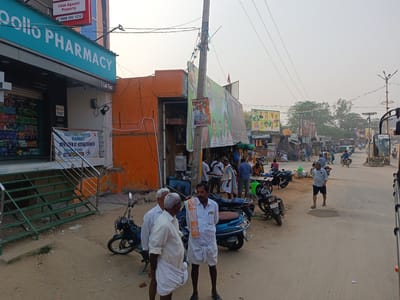 The image depicts a street scene in Kothacheruvu, India. In the foreground, three men in traditional white clothing stand near parked motorcycles. Behind them, a group of people gather near an orange building and a sign with political imagery. The street stretches into the distance with motorbikes, pedestrians, and shops on either side. An Apollo Pharmacy is visible on the left with loan advertisements. Overhead power lines crisscross the sky.